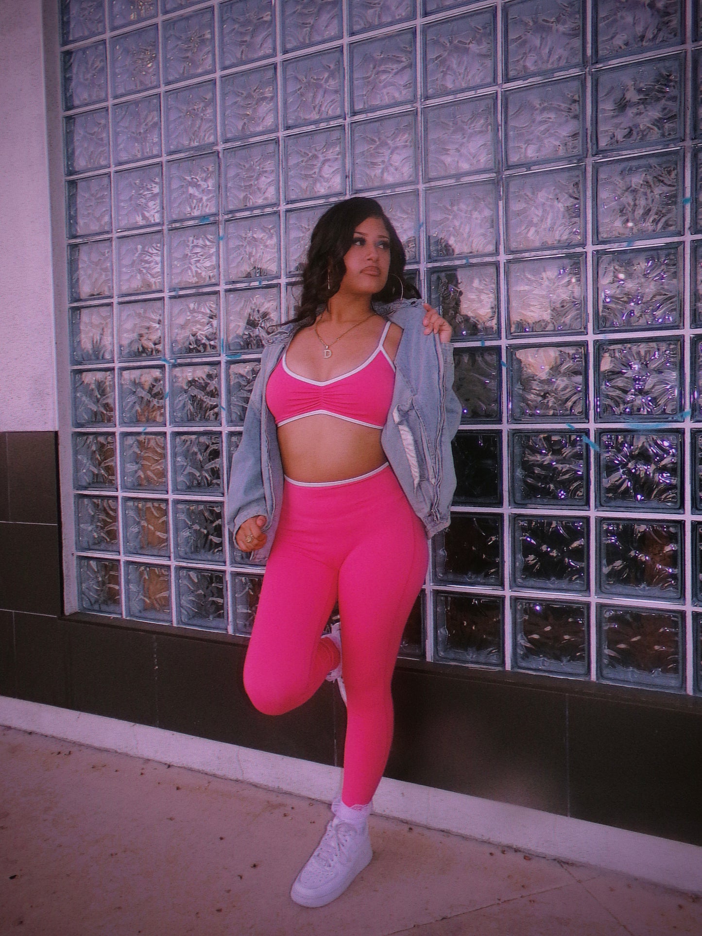 A woman posing in a bright pink activewear set consisting of a sports bra and leggings, styled with a light blue denim jacket and white sneakers, standing in front of a glass block wall. 80's Chicana style.