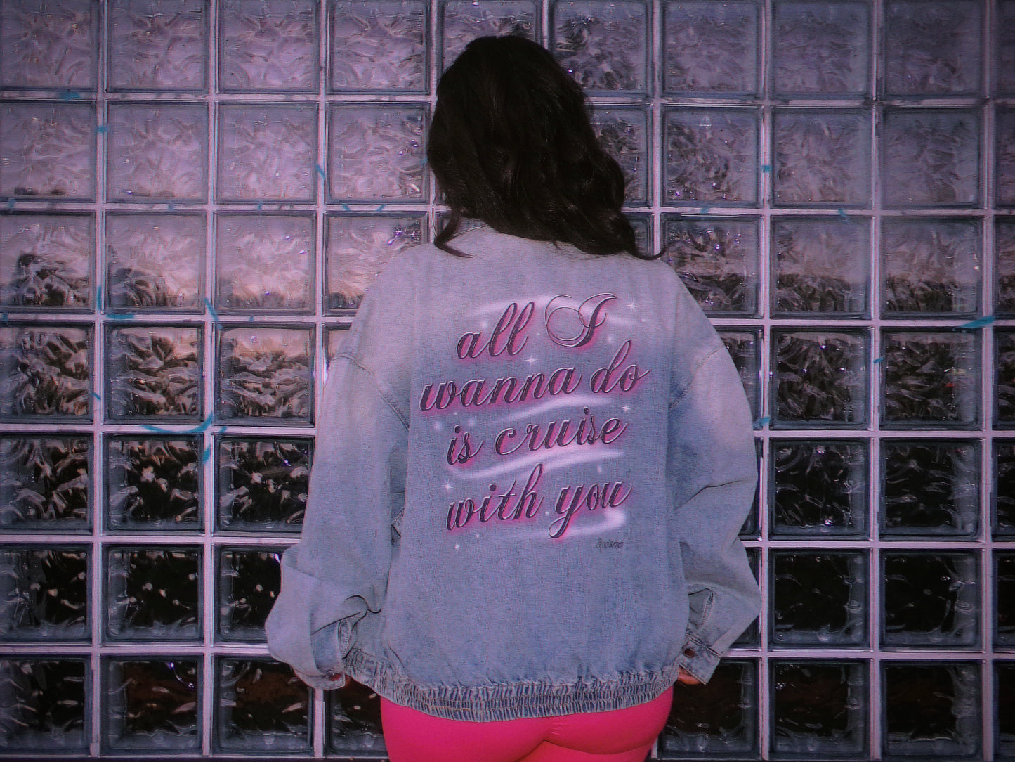 A person viewed from behind wearing a light blue denim jacket with pink airbrush text on the back reading 'all I wanna do is cruise with you', standing in front of a glass block wall. 80's Chicana style.