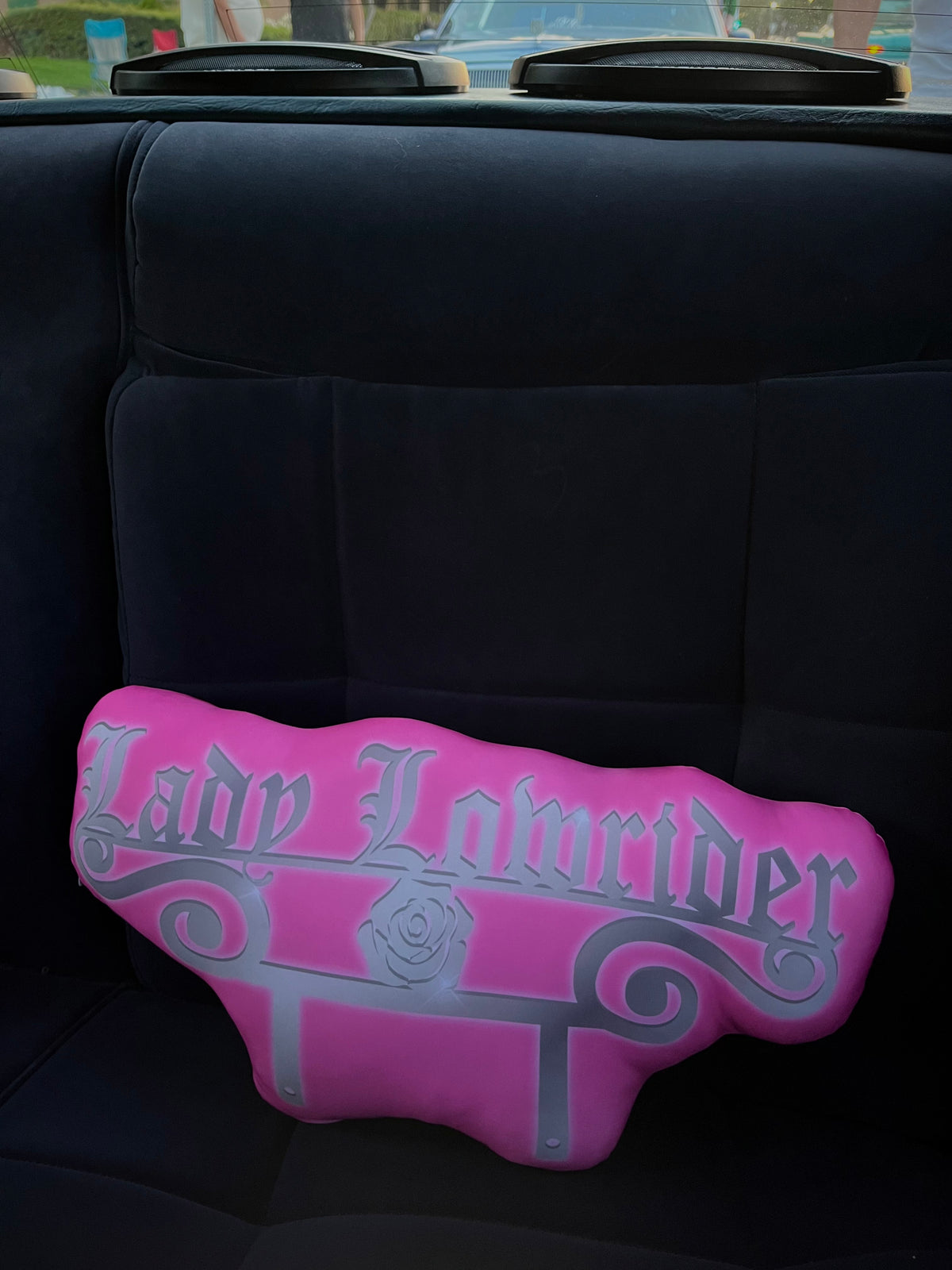 A pink car club plaque pillow with 'Lady Lowrider' written in a gothic font, resting on a car seat.