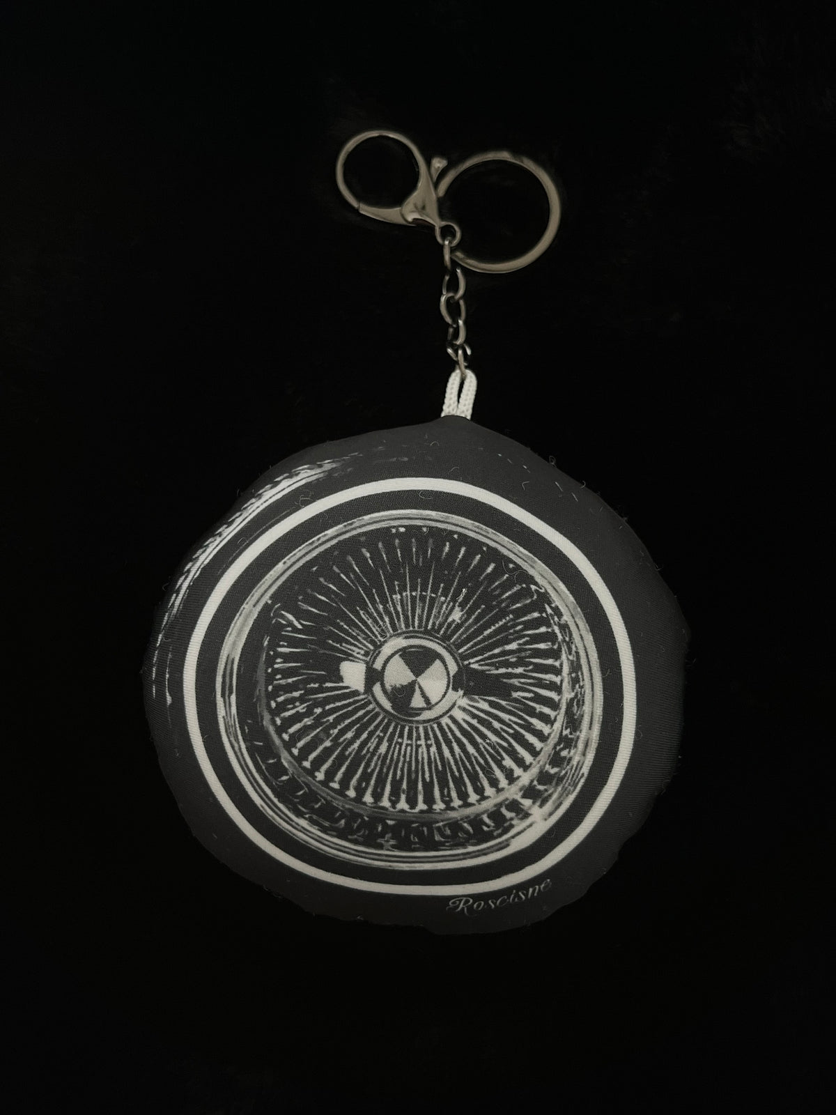 A circular black and white art 100 spoke wire wheel plush keychain with a gunmetal key ring on a solid black background.
