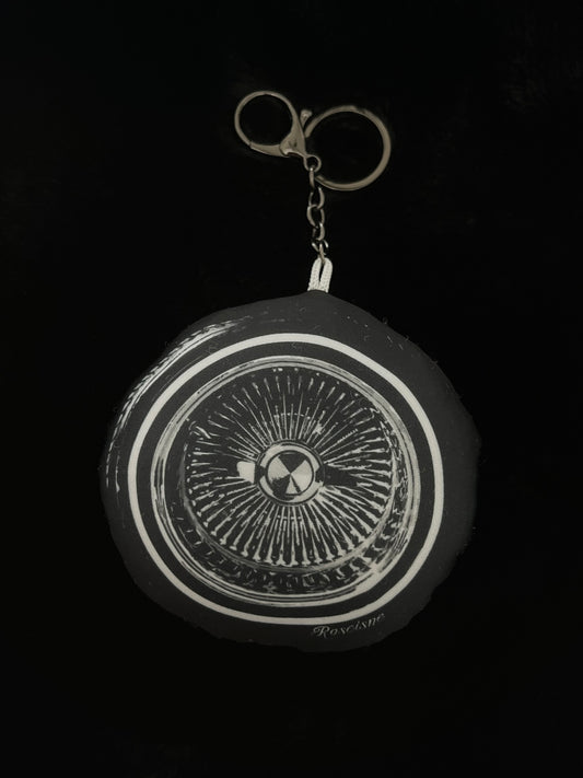 A circular black and white art 100 spoke wire wheel plush keychain with a gunmetal key ring on a solid black background.
