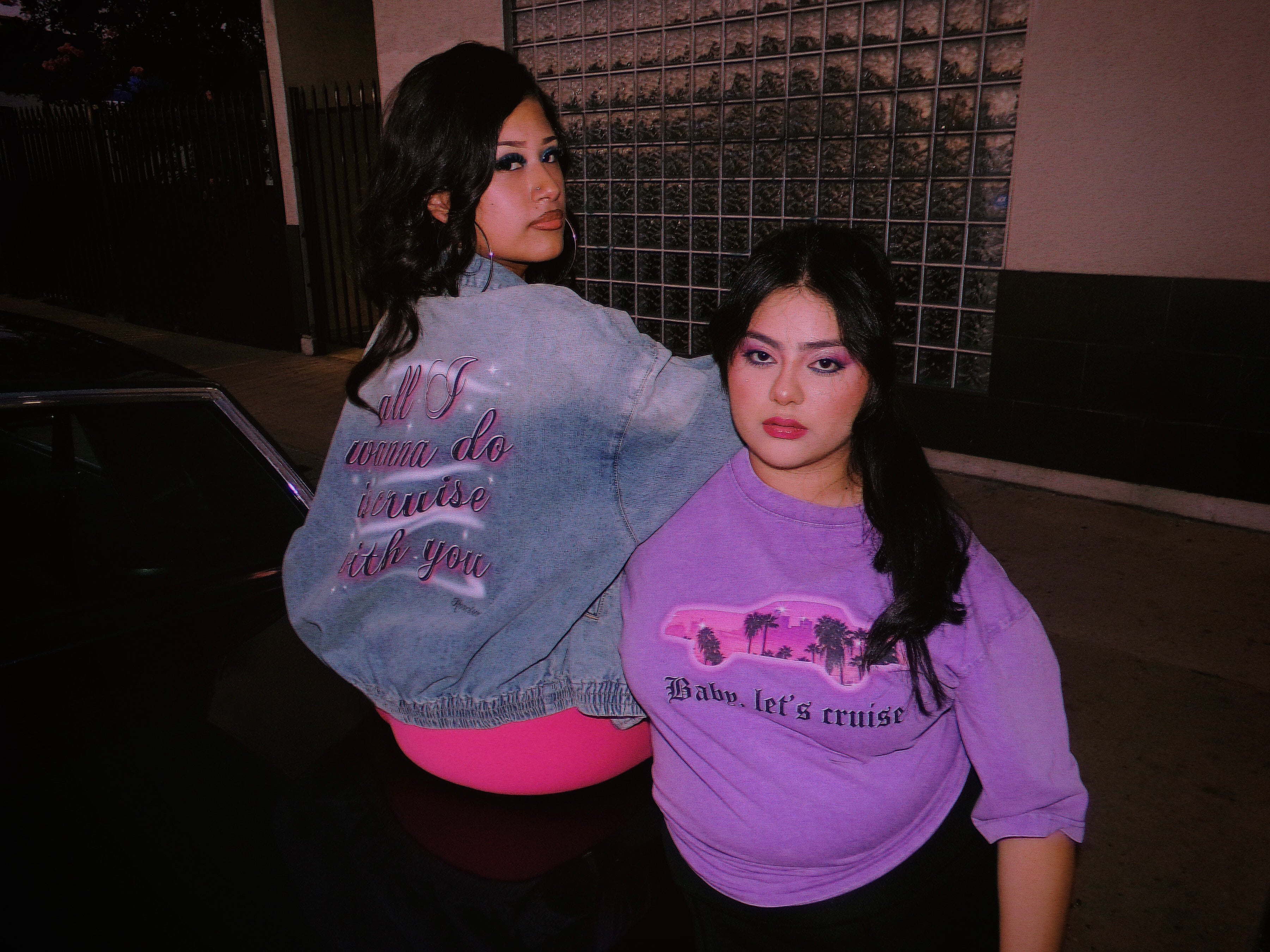 Two women posing outdoors at night under pink-toned lighting on a purple Chevy Monte Carlo lowrider classic car. One wears a light blue denim jacket with pink airbrushed script "all I wanna do is cruise with you' on the back, and the other wears a lavender-purple cropped t-shirt with a pink graphic and the text 'Baby, let's cruise'.