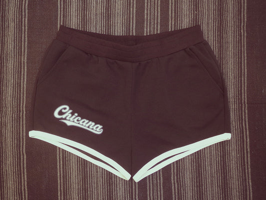 Black 70's style athletic shorts with white trim along the hem and a white script logo reading 'Chicana' on the left leg, laid flat on a serape background.
