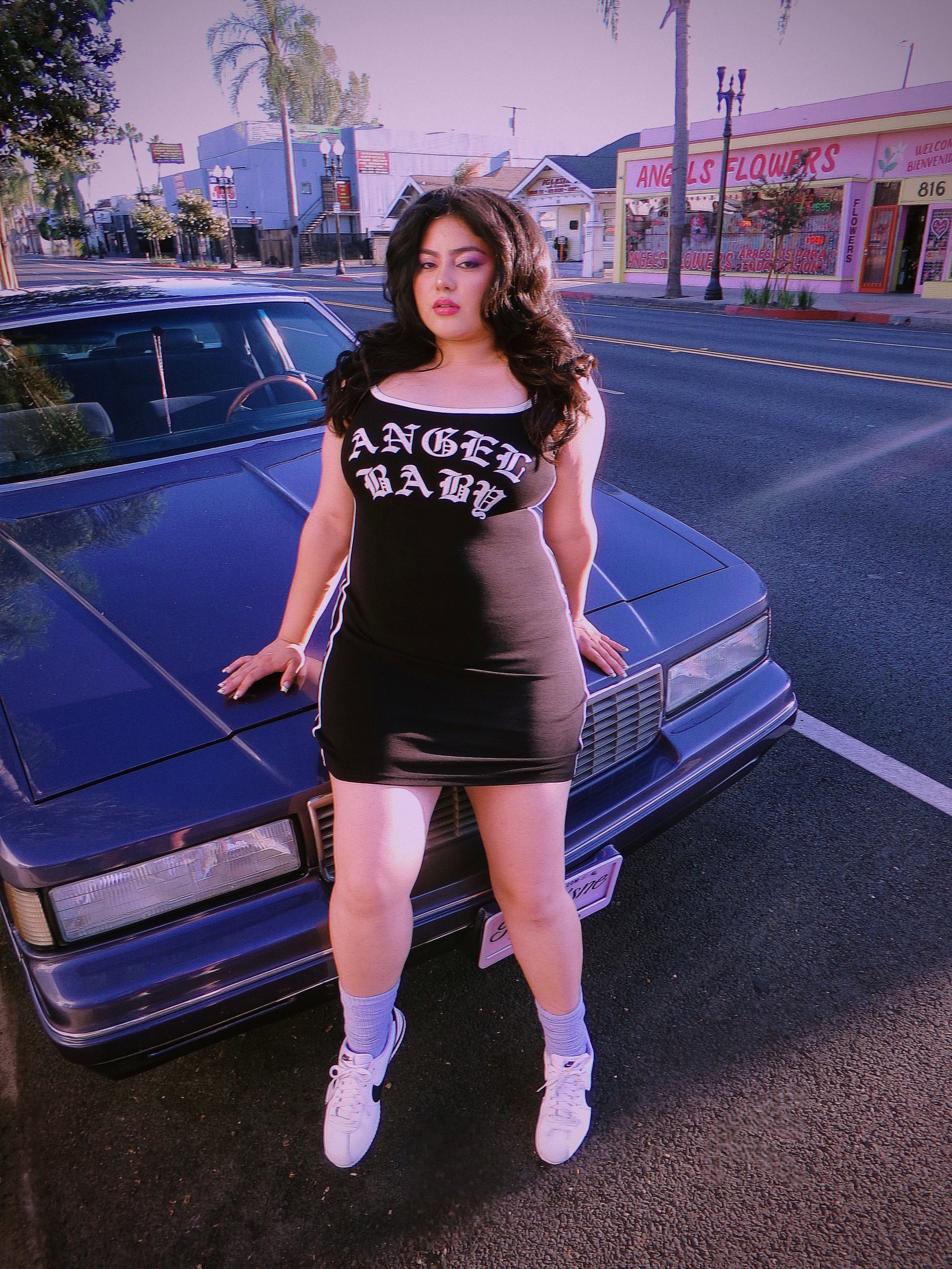 A woman posing in front of a purple lowrider classic car on a street, wearing a tight black mini dress with 'ANGEL BABY' printed in white gothic letters across the chest. 80'c chicana style.