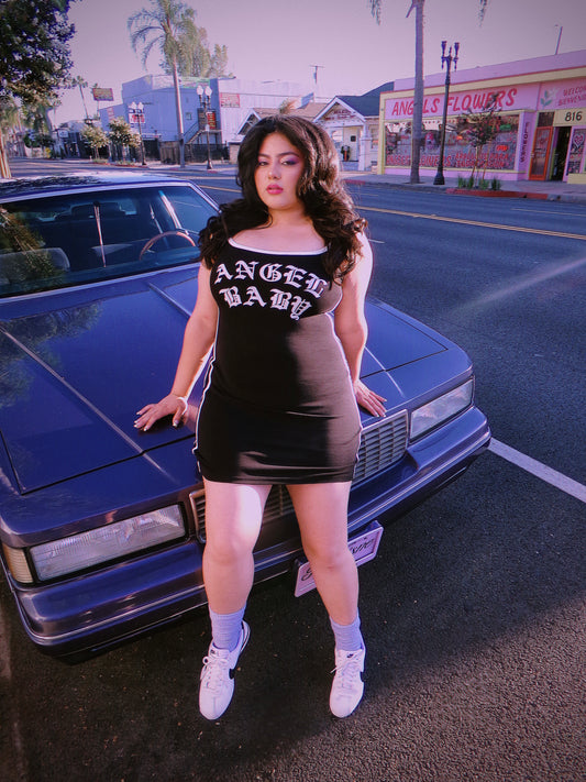 A woman posing in front of a purple lowrider classic car on a street, wearing a tight black mini dress with 'ANGEL BABY' printed in white gothic letters across the chest. 80'c chicana style.