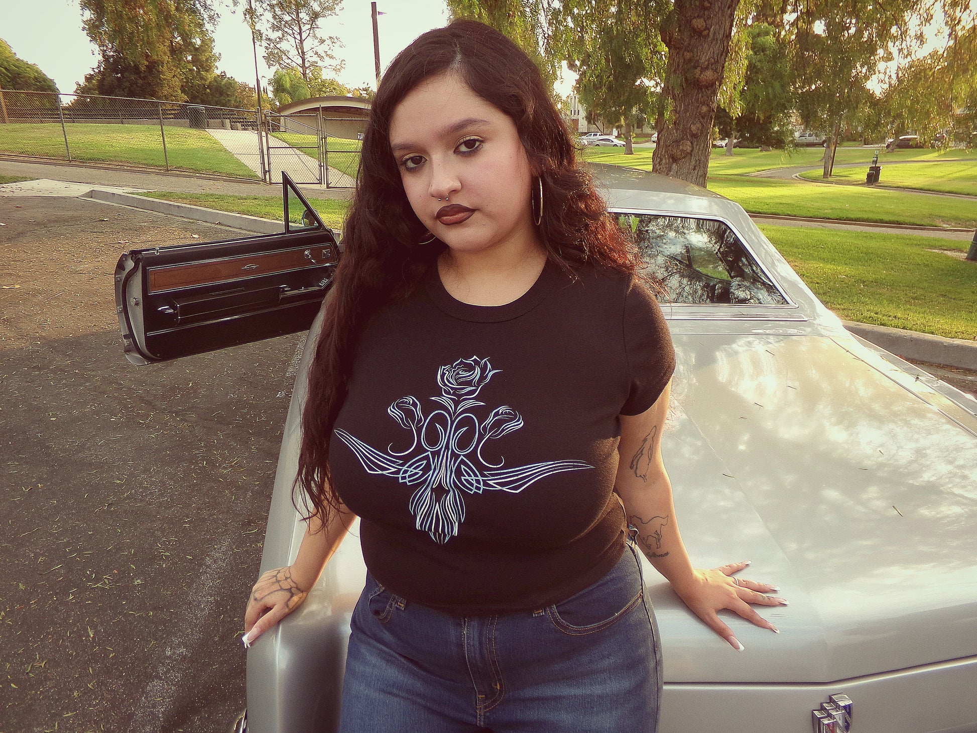 Chicana model at Montebello City park in California next to a silver lowrider car from Lifestyle Car Club, wearing a black short-sleeve t-shirt with a white rose pinstripe graphic.