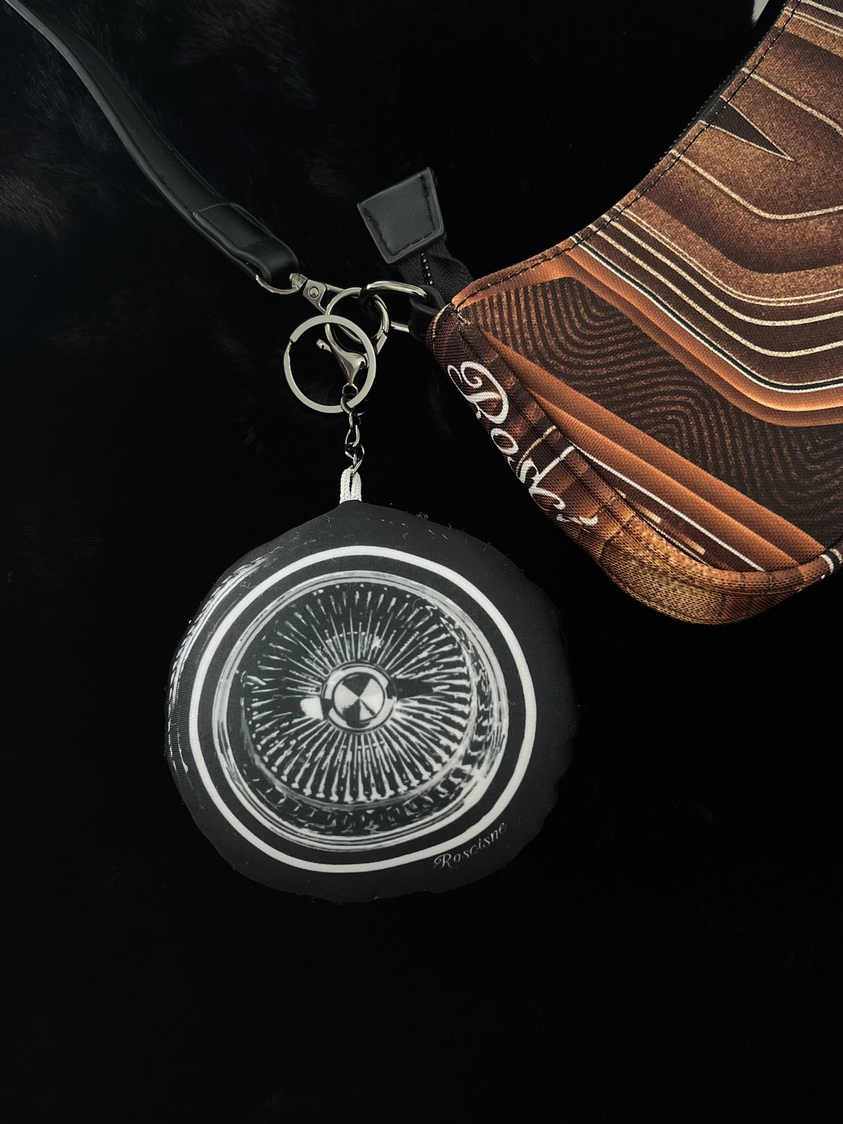 A close-up product shot showing a black circular 100 spoke wire wheel keychain next to a brown lowrider pinstripe patterned purse on a black background.