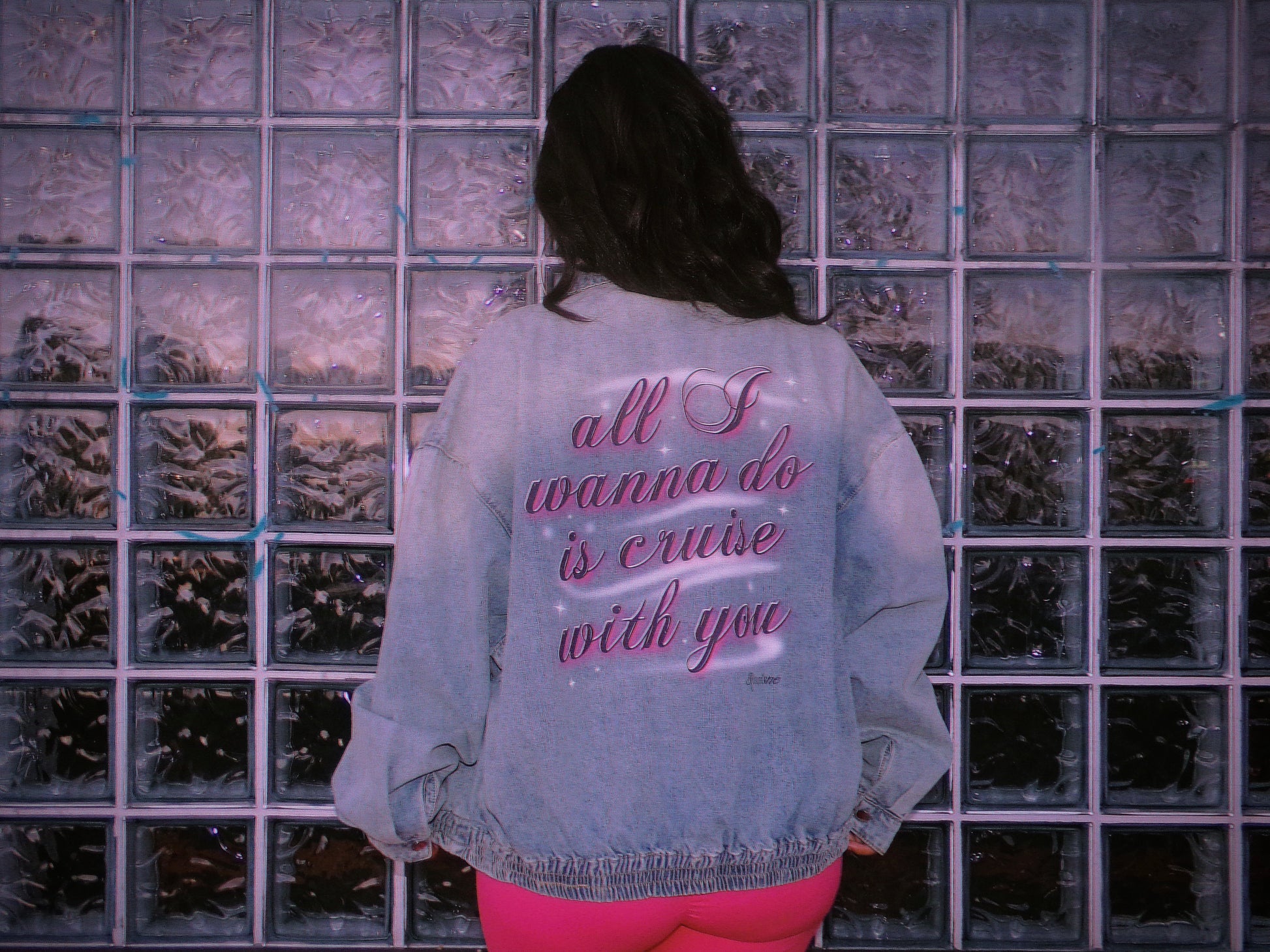 A person viewed from behind wearing a light blue denim jacket with pink airbrush text on the back reading 'all I wanna do is cruise with you', standing in front of a glass block wall. 80's Chicana style.
