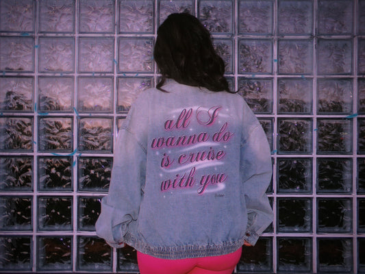 A person viewed from behind wearing a light blue denim jacket with pink airbrush text on the back reading 'all I wanna do is cruise with you', standing in front of a glass block wall. 80's Chicana style.