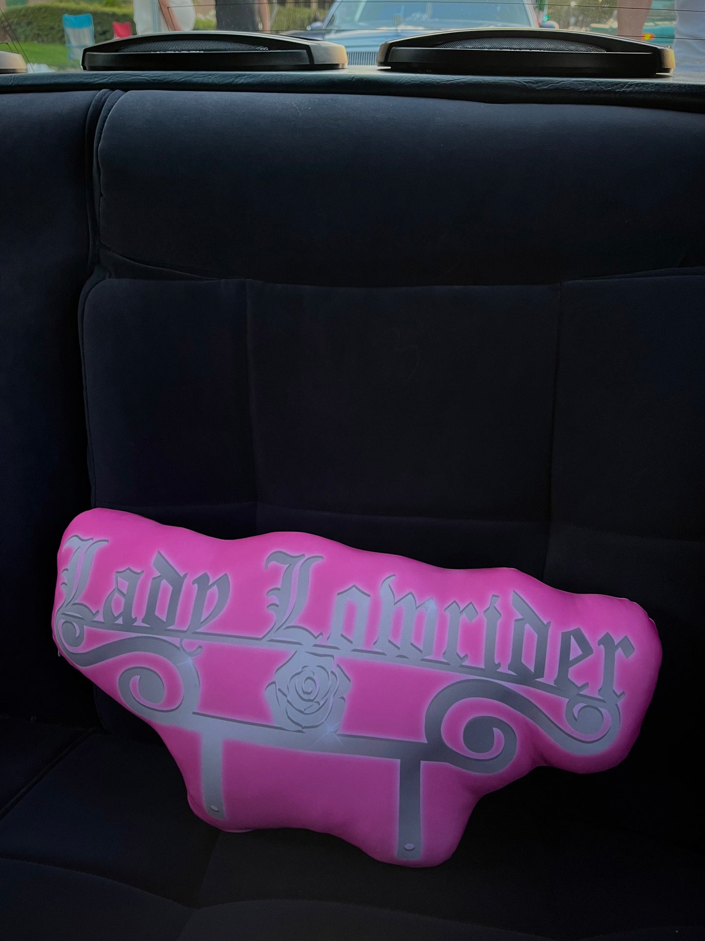 A pink car club plaque pillow with 'Lady Lowrider' written in a gothic font, resting on a car seat.