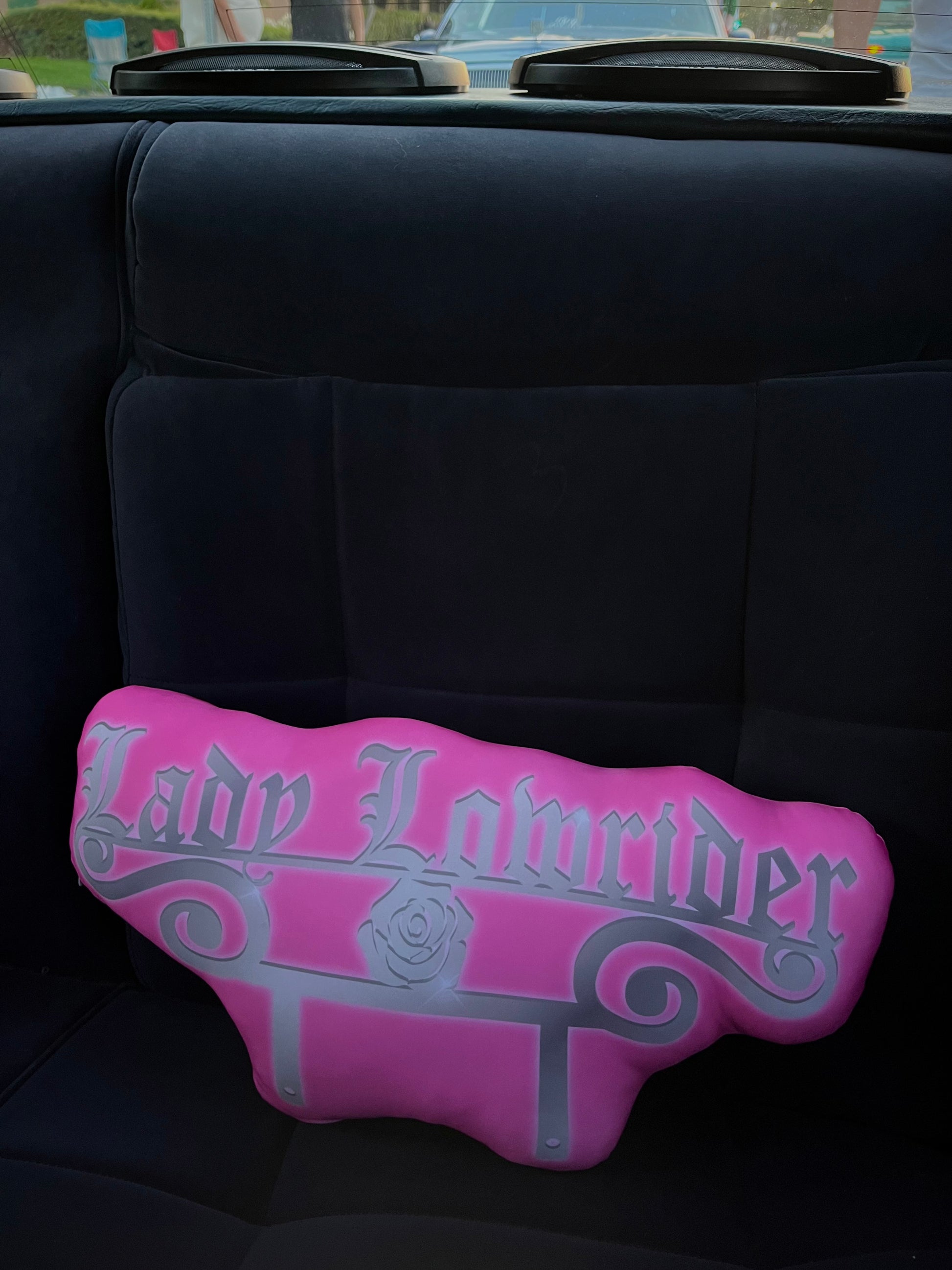 A pink car club plaque pillow with 'Lady Lowrider' written in a gothic font, resting on a car seat.
