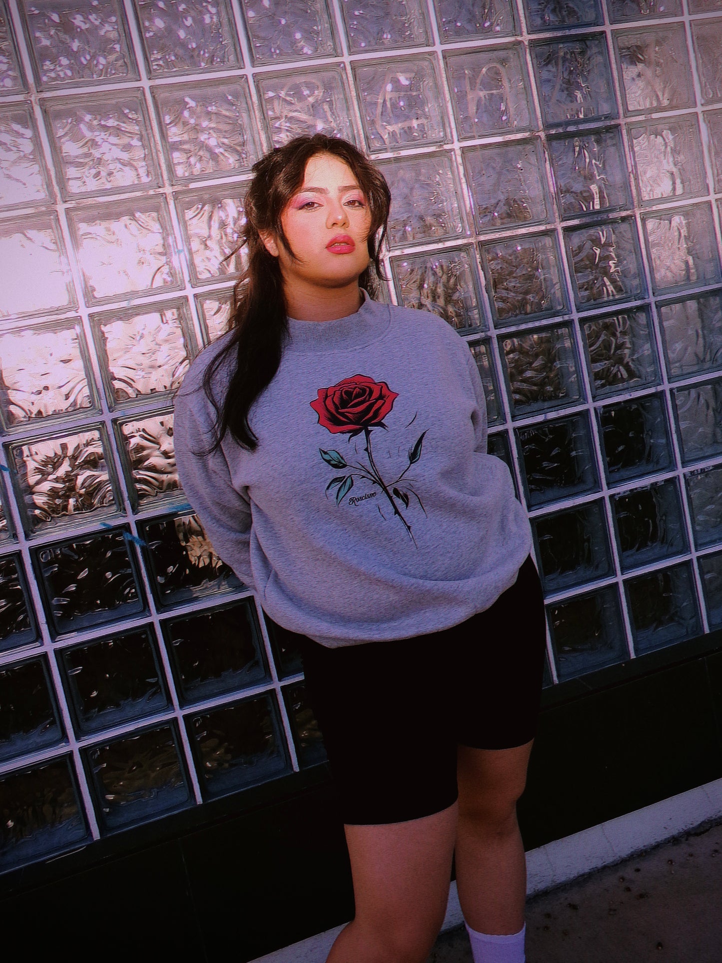 A model wearing a light gray crewneck sweatshirt with a Mexican pink rose graphic on the front, paired with black shorts, standing in front of a glass block wall. 80's Chicana style.